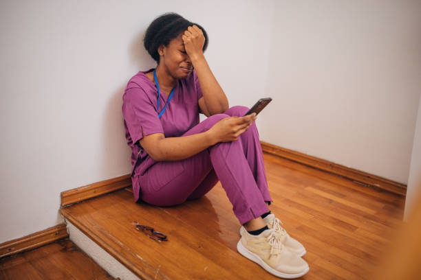 Una mujer con uniforme de trabajo está sentada en el suelo de una habitación con el móvil en la mano y expresión de angustia.
