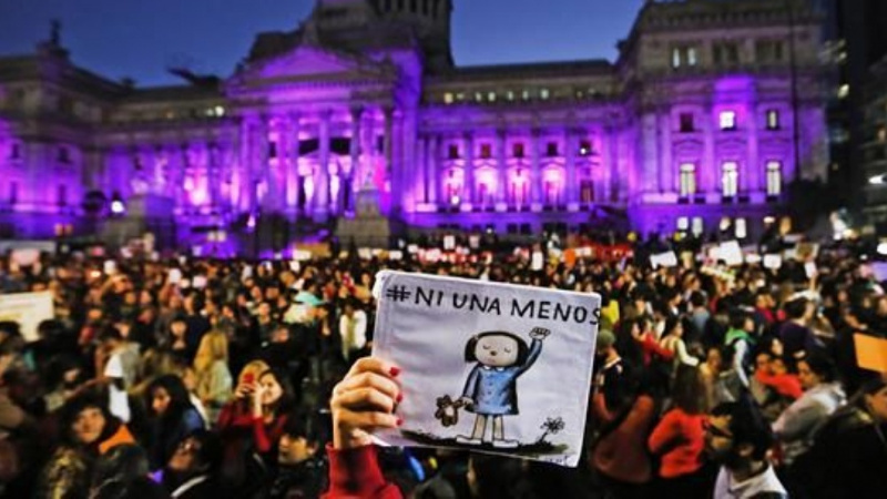 Una mano elevada muestra un dibujo de una niña bajo el hastag #ni una menos, sobre el fondo de un manifestación ante un edificio oficial iluminado con luz morada